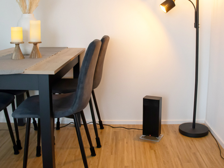 Termowentylator Stadler Form Anna stoi w rogu pokoju obok lampy i stołu z krzesłami.